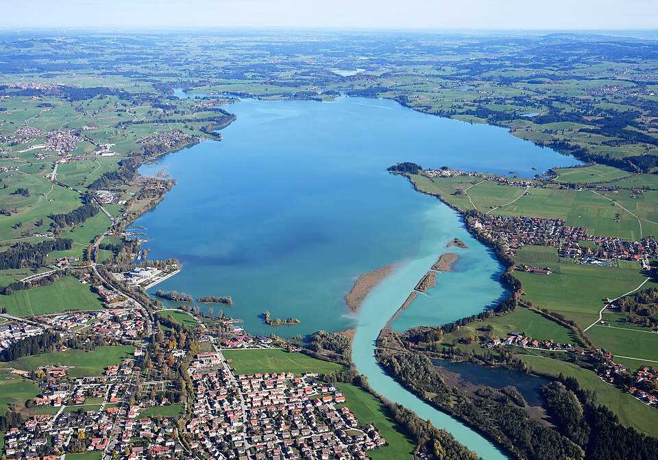 Aerial view of the long Forggensee reservoir with villages and green Allgäu fields around it.