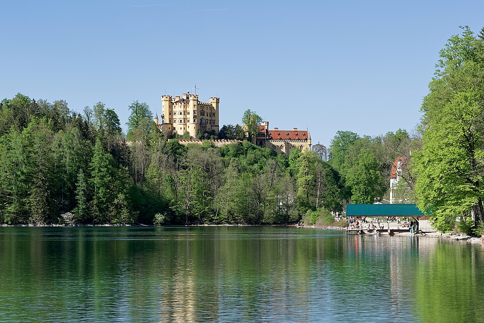 The yellow towers of Schloss Hohenschwangau rising over trees beside the calm Alpsee.